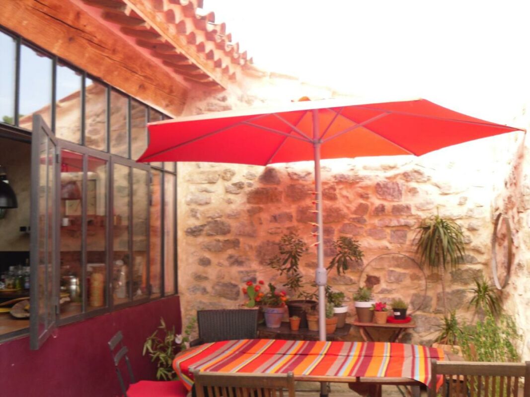 Terrasse en pierre avec table de jardin colorée et parasol rouge, offrant un espace de détente en plein air dans le loft écologique de Villesèque-des-Corbières.