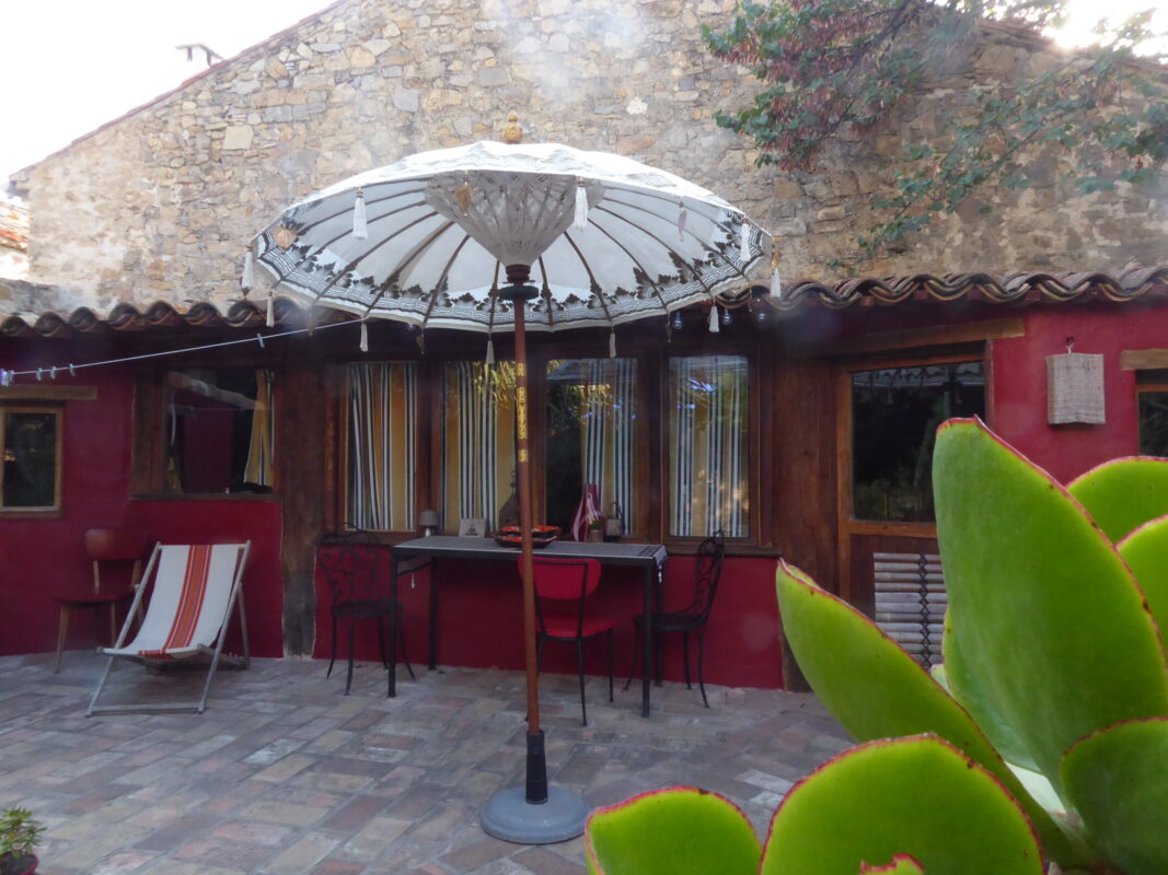 Terrasse ombragée avec parasol et chaises longues, située devant une maison d'hôtes en pierre à Villesèque-des-Corbières.