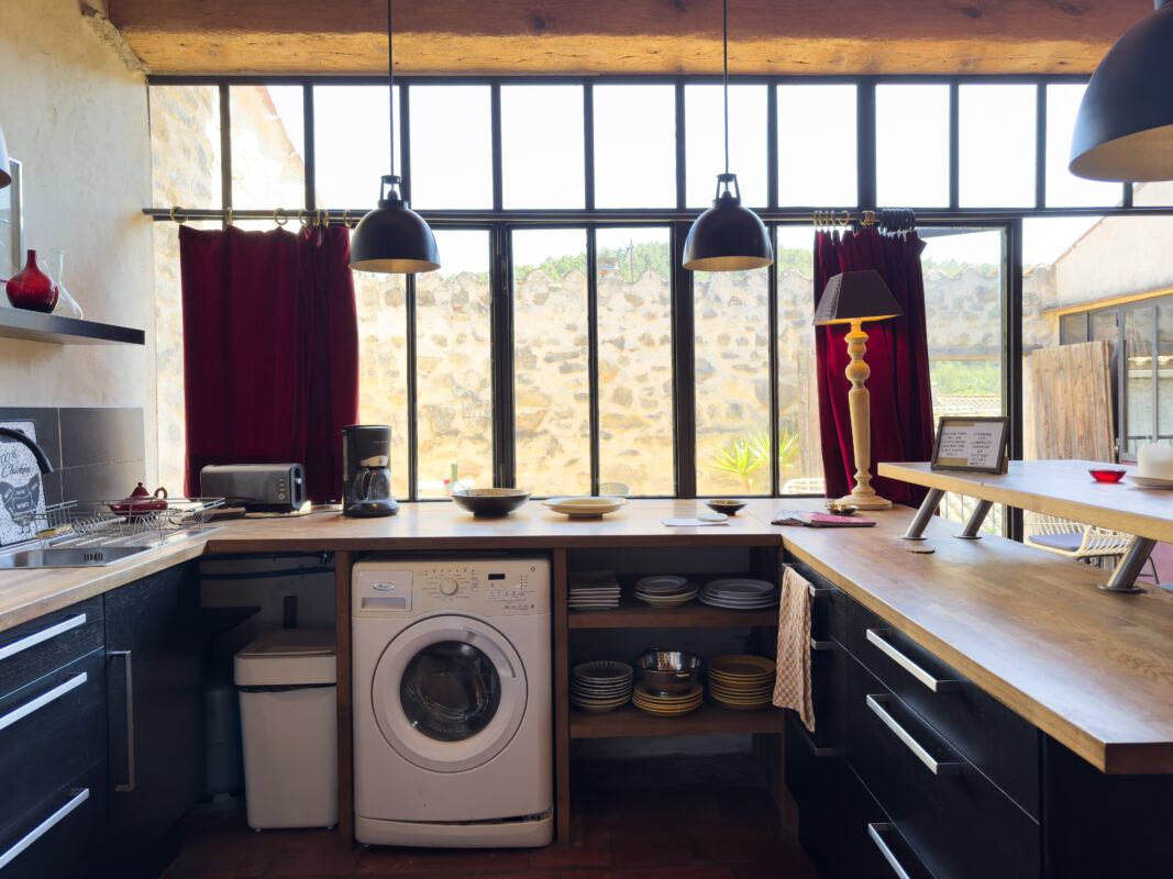 Cuisine lumineuse et rustique de Côté Corbières avec des éléments en bois, un grand plan de travail, une machine à laver intégrée, et de grandes fenêtres donnant sur un mur en pierre.